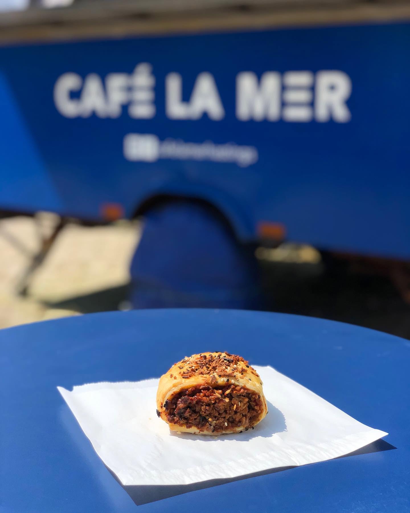 A complete golden savoury pastry on a paper napkin on a blue table, with the Café La Mer trailer softly blurred behind
