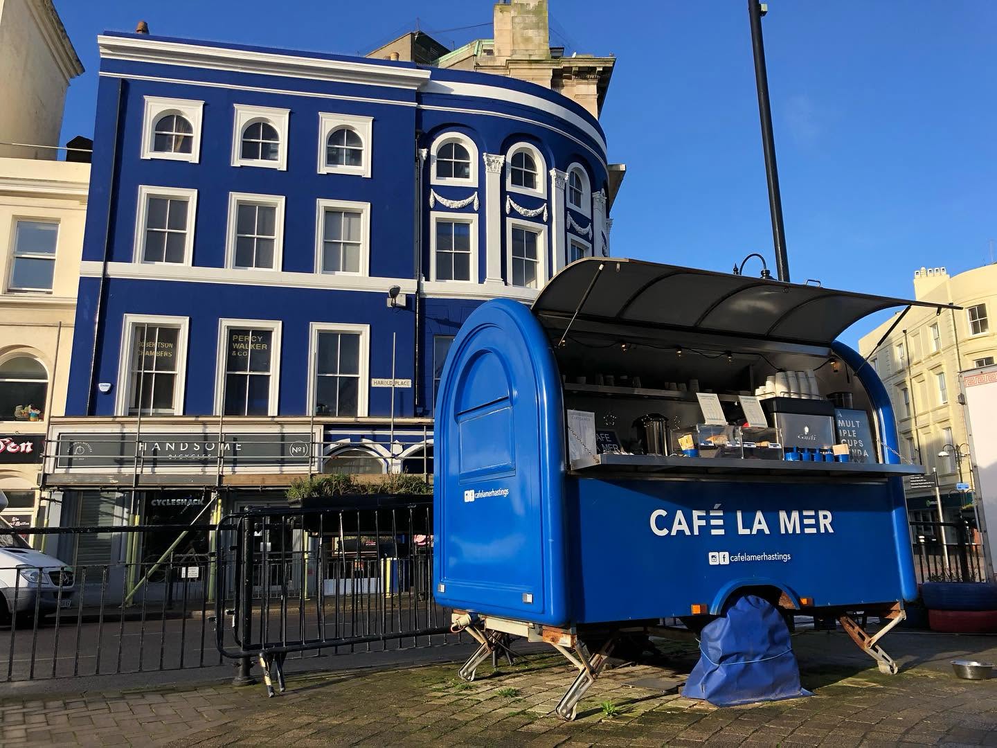 The complete blue Café La Mer coffee trailer with its open service hatch, name, supports and covered wheel in Hastings